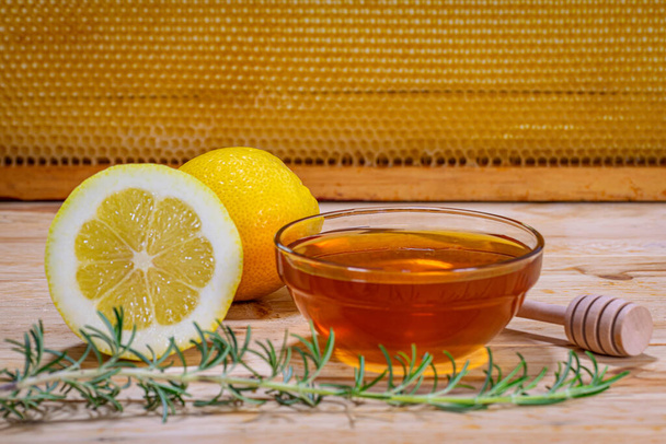 Un tazón con miel, dispensador, una ramita de romero y un limón y medio en madera con un verdadero panal de abeja en el fondo - Foto, imagen