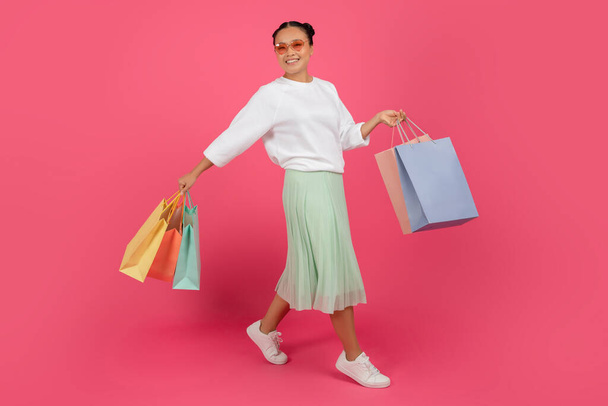 Έννοια αγορών. Ευτυχισμένη όμορφη Ασιάτισσα γυναίκα περπατώντας με φωτεινά τσάντες Shopper σε χέρια πάνω από ροζ φόντο στούντιο, χαρούμενη Shopaholic γυναίκα απολαμβάνοντας εποχιακές εκπτώσεις και μεγάλες πωλήσεις, αντίγραφο χώρου - Φωτογραφία, εικόνα