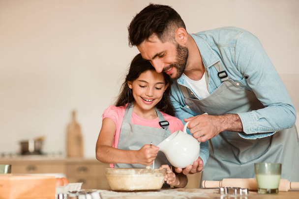 Otec jemně nalévá mléko do misky, zatímco jeho usmívající se dcera míchá těsto, vychutnává si vzácný okamžik sounáležitosti v jejich slunné, domácké kuchyni - Fotografie, Obrázek