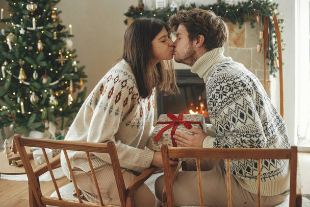 暖炉の背景にスタイリッシュなクリスマスプレゼントを交換する居心地の良いセーターの幸せな若い家族は,ライトでモダンのお祝いのマントルとクリスマスツリー. ハッピー ホリデー! - 写真・画像