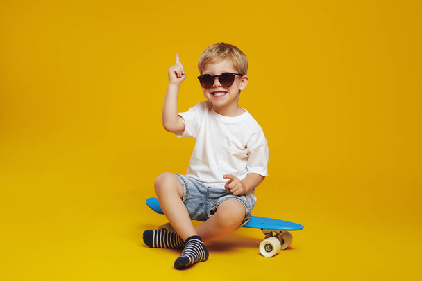 Adorable little boy in white tshirt and sunglasses, sitting on modern skateboard while looking at camera and pointing up, against yellow background. - Photo, Image