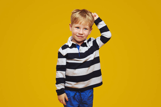 Nachdenkliches kleines Kind mit verwirrter Mimik auf dem Kopf beim Wegschauen, isoliert vor gelbem Hintergrund, in gestreiftem Hemd und blauer Hose. - Foto, Bild