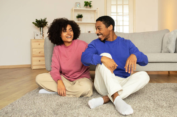 Люблячий чорний чоловік і дружина зв'язуються через ніжні обійми, насолоджуючись романтичними моментами і прихильністю в комфорті свого домашнього простору, сидячи на підлозі разом у інтер'єрі вітальні - Фото, зображення