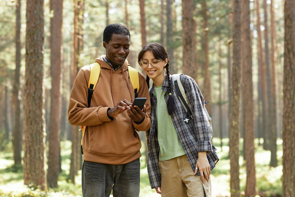 Pareja joven multiétnica usando teléfono móvil mientras está de pie en el bosque durante el senderismo - Foto, imagen