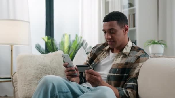 Happy African American man at home couch sofa guy user buyer male client banking buying shopping online with credit bank card and mobile phone smartphone e-commerce order delivery payment internet pay - Footage, Video
