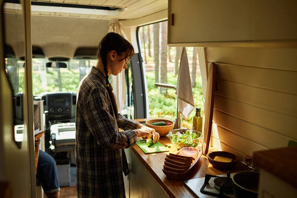 Mujer joven cortando ensalada de verduras en la mesa de la cocina en autocaravana en el camping en el bosque - Foto, imagen