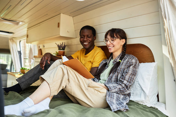 Happy multiethnic couple sitting on bed in house of wheels and reading book together - Photo, Image