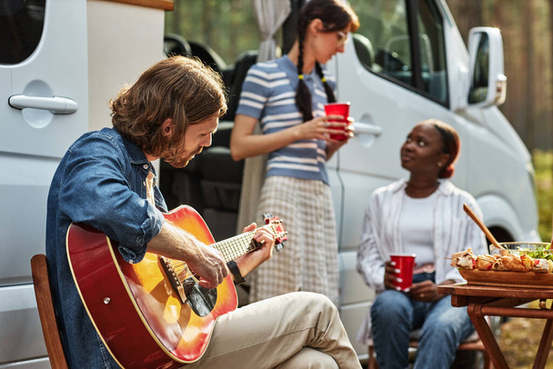 椅子に座って,自然の中でキャンプ中にギターを弾く若い男,2人の女の子が背景で話している - 写真・画像