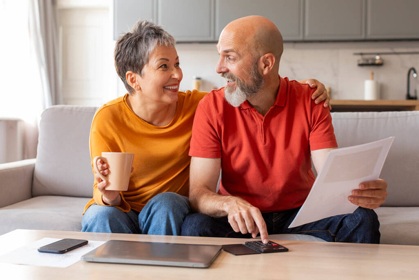 Ευτυχείς ηλικιωμένοι σύζυγοι που ελέγχουν τα οικονομικά έγγραφα και υπολογίζοντας τον οικογενειακό προϋπολογισμό στο σπίτι, χαρούμενο ηλικιωμένο ζευγάρι που κάθεται στο τραπέζι στο σαλόνι, διαβάζοντας τα έγγραφα δανείου ή μετρώντας τις μηνιαίες δαπάνες - Φωτογραφία, εικόνα