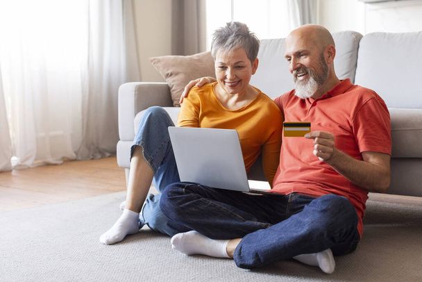 Elderly couple shopping online with credit card and laptop at home, happy senior spouses sitting on floor in living room, retired man and woman using internet banking or making purchases, copy space - Photo, image
