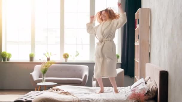 Home ochtend feest in slaapkamer vrolijke vrouw hebben plezier zorgeloos Kaukasisch gelukkig actief meisje in badjas dansen springen op bed bewegen luisteren naar muziek dans springen vreugde verplaatsen vakantie in luxe hotelkamer - Video