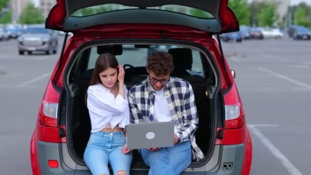 Happy young people sitting on the trunk car outdoors while using laptop, looking on screen. Transportation, technology, trip, travel and lifestyle concept. Real time - Footage, Video