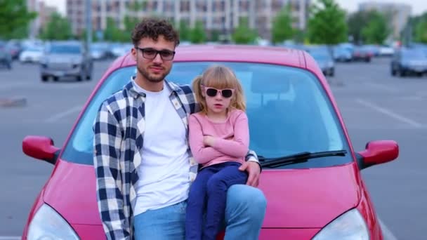 Mosolygó fiatal apa és lánya napszemüvegben hajolt motorháztető autó a szabadban, míg a férfi mutatja hüvelykujját felfelé gesztus. Család, közlekedés, utazás, biztonság és szülőség koncepció. Valós idejű - Felvétel, videó