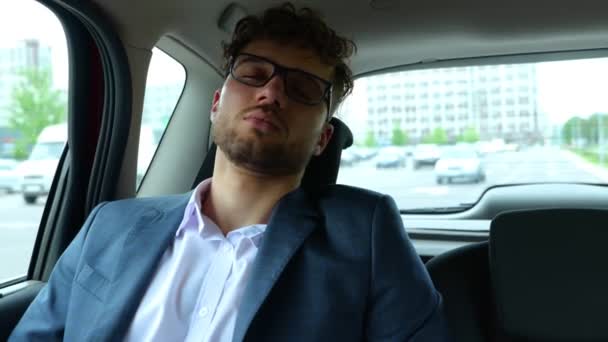 Joven hombre de negocios durmiendo sentado en el asiento trasero del coche, despertando y frotándose los ojos. Concepto de negocios, viajes, transporte y tecnología. Tiempo real - Imágenes, Vídeo