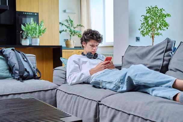 Young handsome guy with headphones using smartphone lying on sofa in living room. Male with curly fashionable hairstyle. Internet online technology mobile applications for study leisure communication - Photo, image