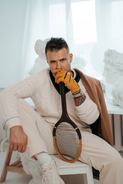 A dapper model in old money style, sitting among ancient Greek sculptures, posing with a tennis racket - Photo, Image