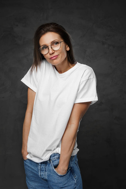 Egy vonzó nő portréja szemüvegben és fehér pólóban, sötét háttérrel. - Fotó, kép