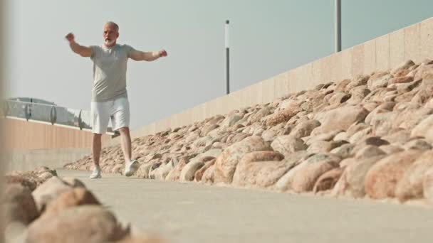 Hombre atlético senior en ropa deportiva caminando hacia la cámara, haciendo círculos de brazos y calentándose antes de la sesión de entrenamiento al aire libre - Metraje, vídeo