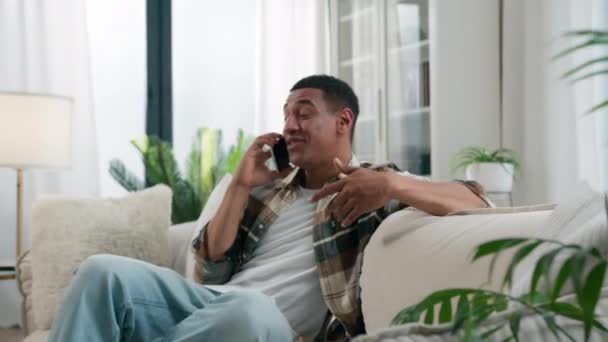 Heureux homme afro-américain à la maison canapé gars mâle insouciant parler smartphone appel avec un ami parler téléphone mobile communication occasionnel agréable parler parler partager nouvelles potins sourire rire - Séquence, vidéo