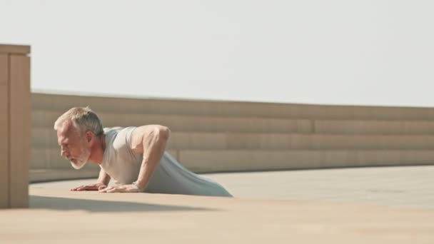 Uomo anziano muscoloso in abbigliamento sportivo che pratica flessioni nello spazio pubblico urbano mentre si allena all'aperto al mattino - Filmati, video