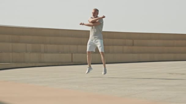 Spor kıyafetlerinde enerjik kıdemli bir adam atlıyor ve dışarıda sabah antrenmanına hazırlanırken omuzlar ve boyun için ısınma egzersizleri yapıyor. - Video, Çekim