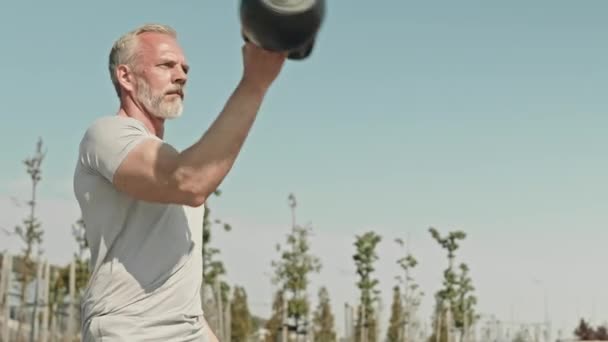 Medium shot of muscular senior man in sportswear doing single arm kettlebell swings and overhead press while exercising outdoors in city - Footage, Video