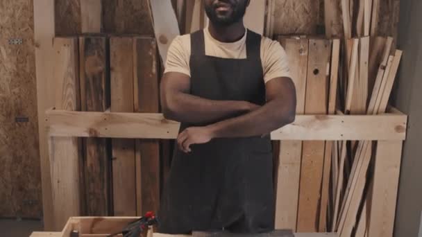 Incline-se retrato de carpinteiro afro-americano alegre em óculos e avental em pé em seu estúdio ao lado da mesa e olhando para a câmera - Filmagem, Vídeo