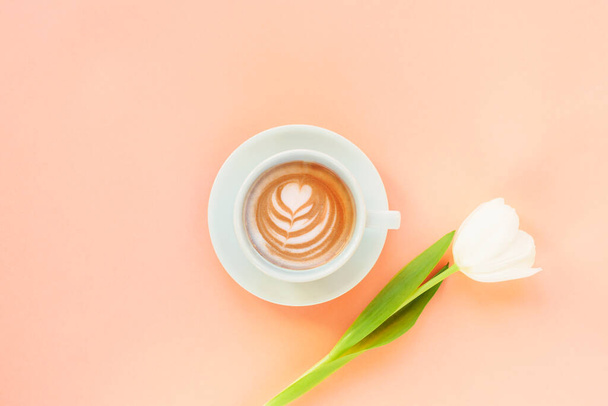 Tasse de café avec art du café et tulipe blanche sur fond de couleur pêche. Journée des femmes ou concept de fête des mères. Vue du dessus, plan, espace de copie. - Photo, image