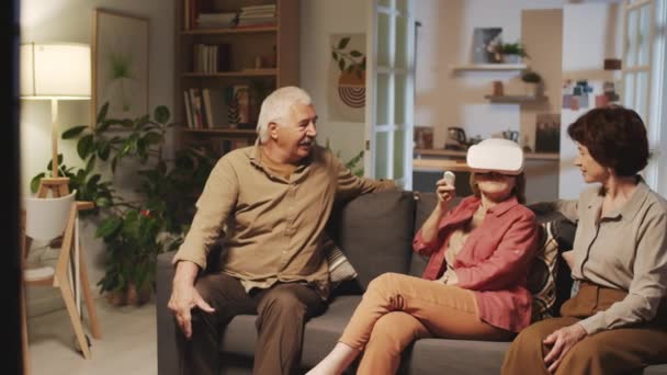 Средний длинный снимок возбужденной пожилой женщины, впервые пытающейся надеть VR-очки, проводящей время на вечеринке с друзьями в уютной гостиной - Кадры, видео