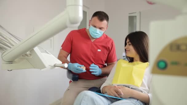 マスクドクター歯科医を身に着けている若い矯正歯科医の男性は歯科オフィスの椅子の室内キャビネットの近くに座っている患者のための歯科プラスチック顎の咬傷を示します.  - 映像、動画
