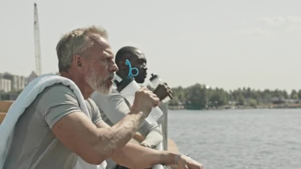 Starszy mężczyzna i młody czarny mężczyzna opierający się na balustradzie na miejskim brzegu rzeki, pijący wodę i rozmawiający podczas odpoczynku po treningu na świeżym powietrzu w letni dzień - Materiał filmowy, wideo