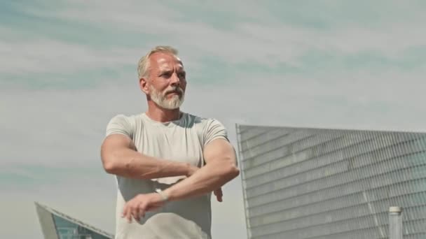 Πορτρέτο του μυώδους ηλικιωμένου άνδρα σε σπορ t-shirt στέκεται σε εξωτερικούς χώρους με τα χέρια σταυρωμένα και ποζάρουν για την κάμερα κατά ουρανοξύστες - Πλάνα, βίντεο