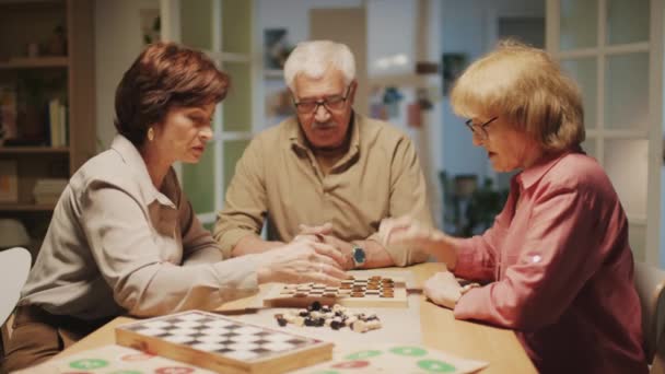 Mediana toma de amigos ancianos jugando damas mientras están sentados en la mesa juntos en casa - Imágenes, Vídeo
