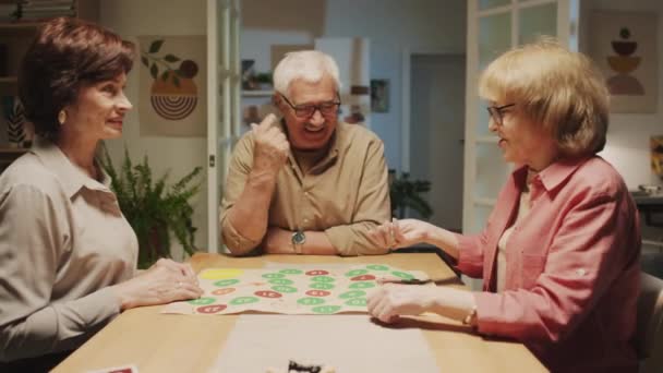 Střední záběr společnosti pozitivních seniorů, kteří hrají deskovou hru a sedí u stolu na domácí párty - Záběry, video