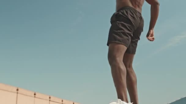Colpo basso angolo di uomo afroamericano senza maglietta che fa squat di salto in panchina mentre si allena all'aperto in città il giorno d'estate - Filmati, video