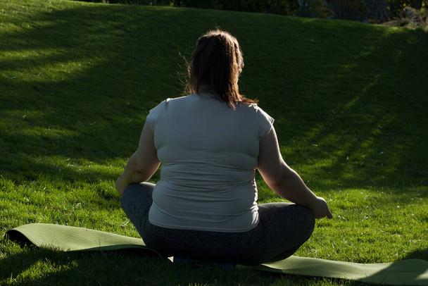 Portrait of overweight Caucasian middle-aged woman practicing yoga in nature. Fat woman meditating, relaxing in lotus position, sitting on fitness mat. Calm, relaxed, zen, stop stress. View from back - Photo, Image