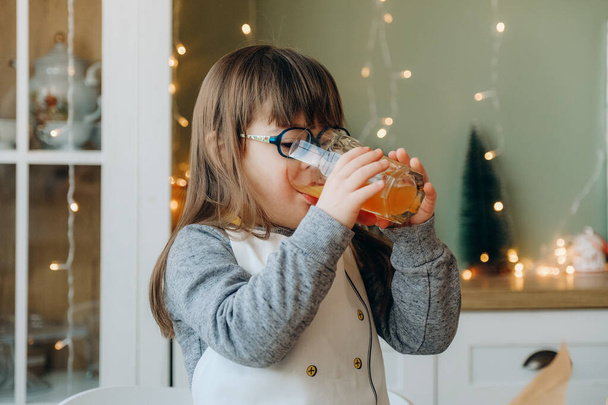 Uma menina com síndrome de Down bebe suco de laranja e se senta na mesa na véspera de Natal. - Foto, Imagem