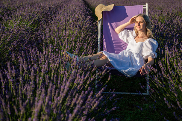 Chica joven y relajada en un vestido blanco, sentada en una silla, respirando aire fresco, sentada en un campo de lavanda en un día soleado, mirando hacia un lado, pensando - Foto, imagen