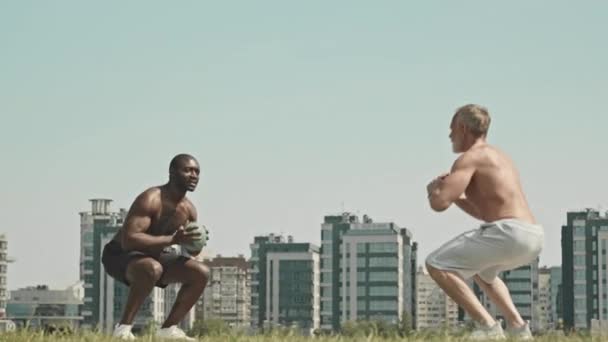 Gros plan de musclé torse nu homme noir et faire partenaire med balle squat jette tout en ayant l'entraînement en plein air avec un ami aîné - Séquence, vidéo
