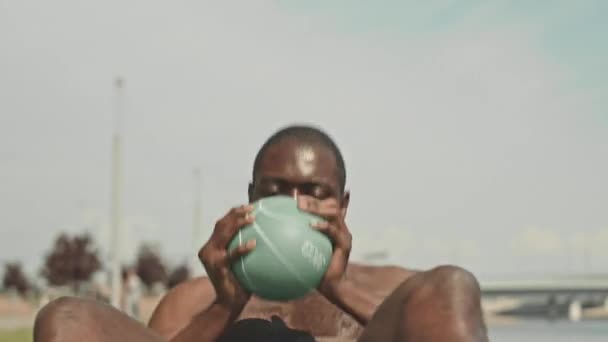 Czarny mężczyzna bez koszuli z muskularnym tułowiem robi brzuszne chrupania i skręty z piłką medyczną podczas treningu na świeżym powietrzu w letni dzień - Materiał filmowy, wideo