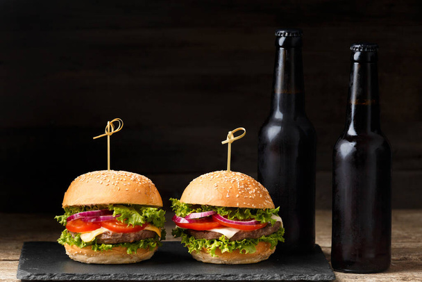 Dwa hamburgery, frytki, piwo w ciemnych butelkach na drewnianym tle. Fast food, domowe hamburgery - Zdjęcie, obraz
