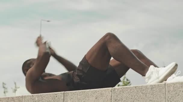 Low angle shot of shirtless African American man doing abdominal crunches while exercising outdoors on summer day - Footage, Video