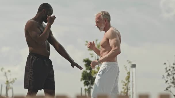 Μεσαία μακριά βολή της μυϊκής Αφρικής Αμερικανός εκπαιδευτής διδασκαλία ανώτερος Καυκάσιος άνθρωπος kickboxing τεχνική κατά τη διάρκεια υπαίθριο μάθημα στο δρόμο - Πλάνα, βίντεο