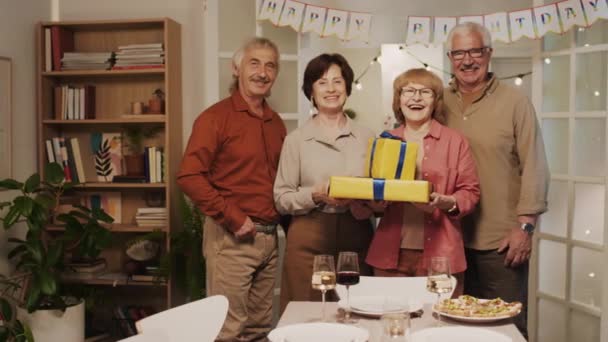 Retrato de amigos ancianos felices de pie en la cómoda sala de estar con regalos y mirando a la cámara en la fiesta de cumpleaños en casa - Metraje, vídeo