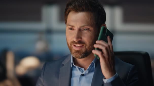Бородатий чоловік відповідає на дзвінок в нічний корпоративний офіс крупним планом. Усміхнений чоловік розмовляє мобільним телефоном, реагуючи на хороші новини в вечірньому інтер'єрі. Позитивний бізнесмен спілкується з другом на темному робочому місці  - Кадри, відео
