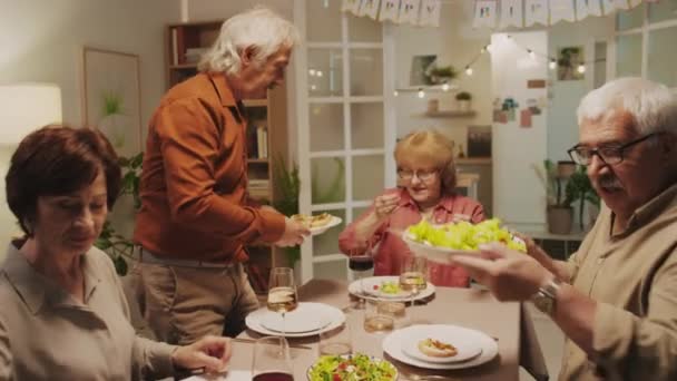 Medium shot of senior couple treating friends with festive dishes sitting together at table at cozy home birthday celebration party - Footage, Video