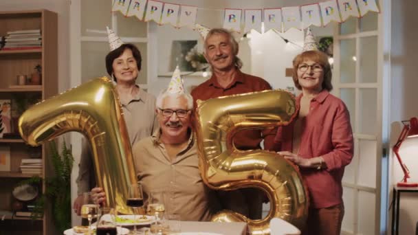 Portrait shot of happy pensioners posing for camera with balloons in shape of number seventy five when celebrating anniversary at home party - Footage, Video