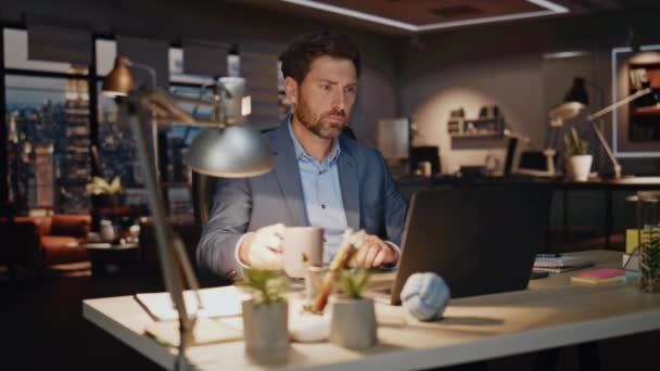 Бізнесмен п'є каву ввечері на робочому місці крупним планом. Думаючи, що бородатий працівник сіє чашку чаю, дивлячись на екран ноутбука. Орієнтований робочий процес вирішення складних завдань друку комп'ютера в нічному кабінеті  - Кадри, відео