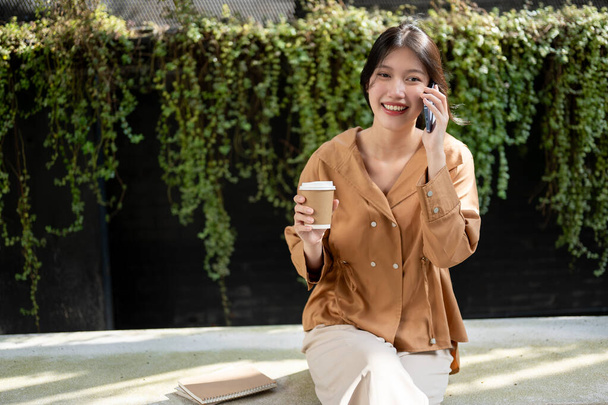 Uma bela e feliz jovem asiática está gostando de falar ao telefone com sua amiga e tomar café enquanto relaxa em um banco no jardim em um dia brilhante. Pessoas e conceitos de estilo de vida - Foto, Imagem