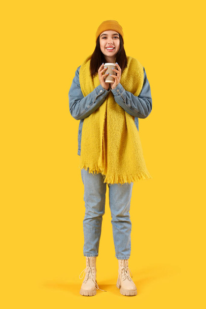 黄色い背景のコーヒーカップと冬の服の若い女性 - 写真・画像